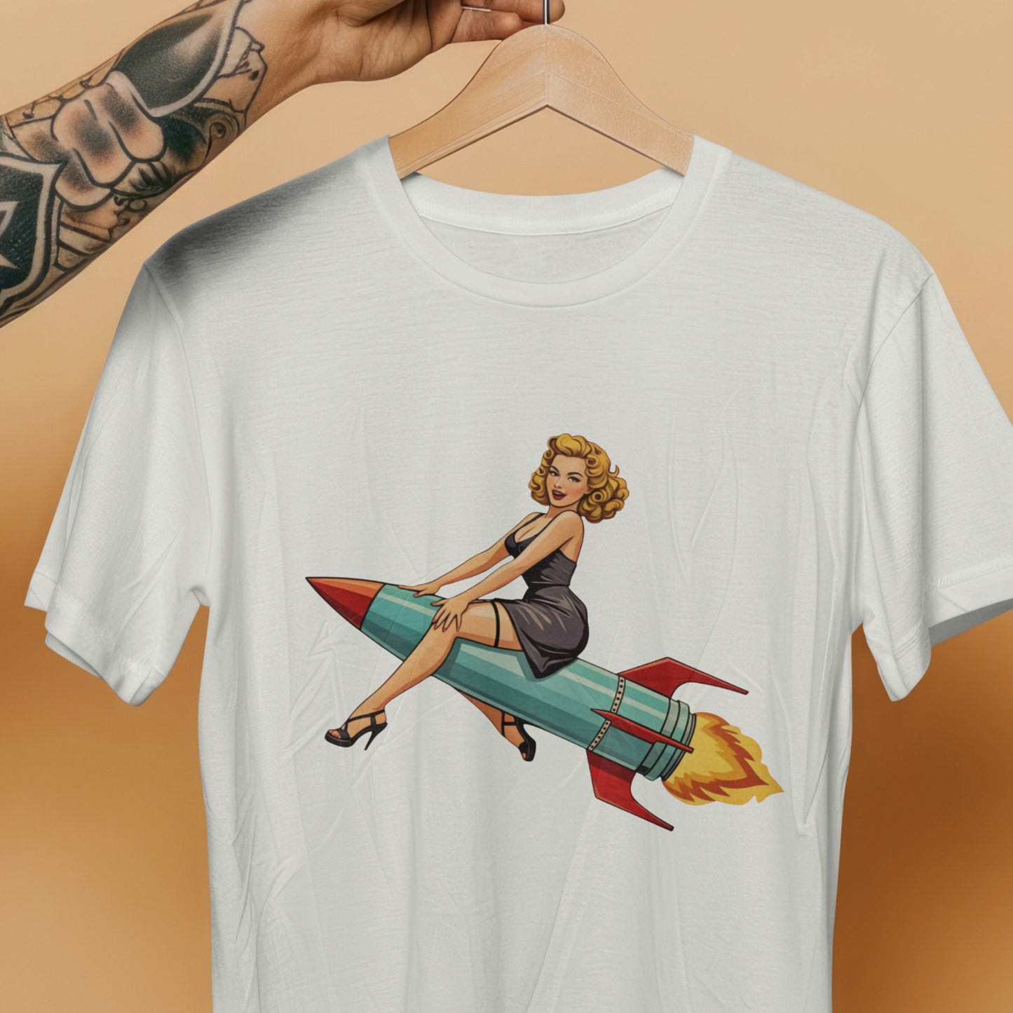 White t-shirt with a graphic of a woman on a rocket, hanging on a wooden hanger against a beige background.